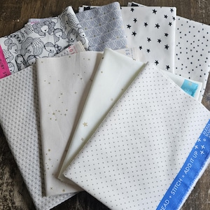 Op de afbeelding: Een stapel van acht stofvierkanten met verschillende patronen. De patronen omvatten zwart-witte geometrische ontwerpen, zwarte sterren op een witte achtergrond en gouden sterren op een witte achtergrond. De stofvierkanten zijn allemaal wit met verschillende patronen.