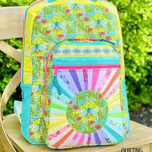Può includere: Uno zaino colorato con un vivace motivo a api e fiori. La tasca anteriore presenta un design a raggi di sole arcobaleno con un motivo circolare di api e foglie. Lo zaino è realizzato in tessuto trapuntato.