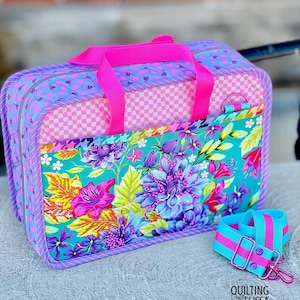 May include: A rectangular quilted fabric bag featuring a vibrant floral pattern in teal, purple, and pink, with a pink checkered panel, bright pink nylon handles, and a detachable matching shoulder strap. The text QUILTING ELISSA is visible.