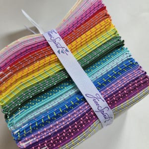 Puede incluir: Un conjunto de cuadrados de tela coloridos en degradado arcoíris, atados con una cinta blanca impresa con "Free Spirit". La tela presenta un patrón de puntos en varios colores, creando una apariencia vibrante y texturizada. Ideal para proyectos de patchwork y manualidades.