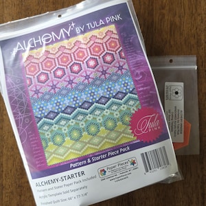 May include: A clear plastic package labelled "ALCHEMY BY TULA PINK" contains a colourful quilt pattern and starter piece pack. The pattern features a geometric design in shades of pink, blue, green, and yellow. A smaller package with an orange piece is also visible.