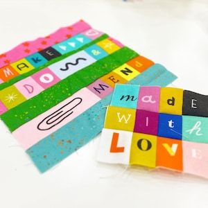 May include: Two fabric patches with text in various colours. One reads "MAKE DO MEND" with a paperclip graphic. The other reads "made with LOVE". The patches feature pink, green, blue, orange, and black colours.