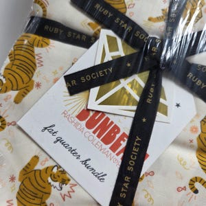 Op de afbeelding: Een stoffen bundel verpakt in doorzichtig plastic, vastgebonden met zwart lint bedrukt met "RUBY STAR SOCIETY" in goud. De stof heeft een herhalend patroon van tijgers in oranje en geel. Een label leest "SUNBEAM" en "fat quarter bundle".