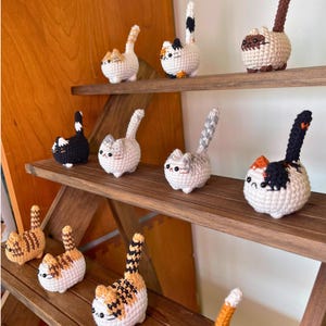 Op de afbeelding: Een verzameling van tien kleine, ronde, gehaakte kattenfiguurtjes op houten planken. Elke kat heeft een uniek kleurpatroon, waaronder oranje, wit, zwart, bruin en grijs, met rechtopstaande staarten, kleine oortjes en geborduurde gezichtskenmerken.
