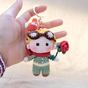May include: A crochet keychain featuring a small doll with yellow hair, brown goggles, a red scarf, and a green outfit. The doll holds a red rose in one hand and has a small orange fox figure perched on its head, all attached to a silver metal ring.