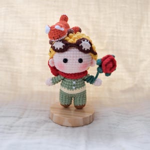 May include: A crocheted doll with yellow hair, brown goggles, and a red scarf, wearing a light green outfit with two white buttons. The doll has a small orange fox on its head and holds a red rose in one hand, all mounted on a round wooden base.