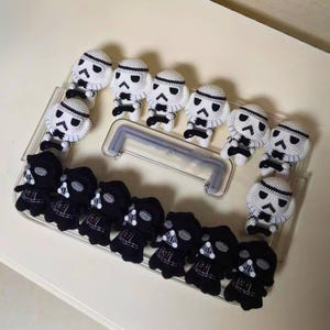 May include: A collection of small, handmade crochet figures arranged on a clear plastic tray. The set includes several white and black stormtrooper characters and black Darth Vader characters, all featuring detailed stitched facial features.