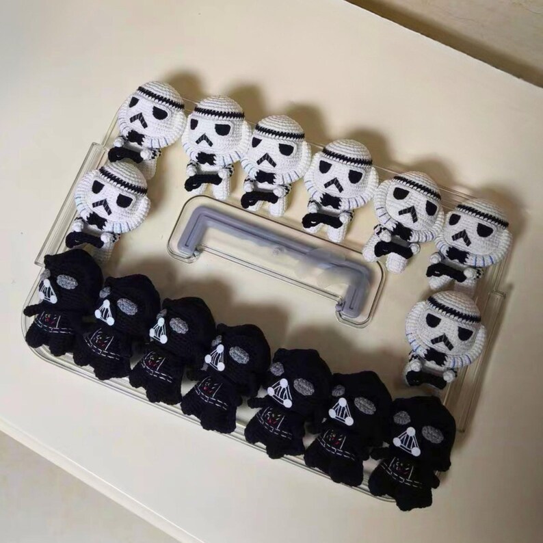 May include: A collection of small, handmade crochet figures arranged on a clear plastic tray. The set includes several white and black stormtrooper characters and black Darth Vader characters, all featuring detailed stitched facial features.
