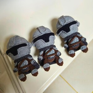 May include: Three handmade crochet dolls shaped like armored warriors. Each doll features a grey helmet with a black visor, a grey torso, and brown accents representing straps and boots. The dolls are crafted with visible yarn stitches.