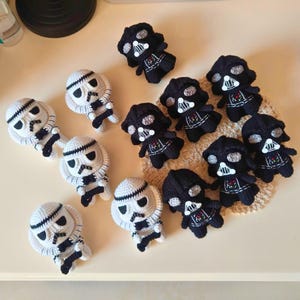 May include: A collection of small, handmade crochet dolls featuring black Darth Vader figures and white Stormtrooper figures. Each character has distinct stitched facial details, including large eyes and mask patterns, crafted with yarn.