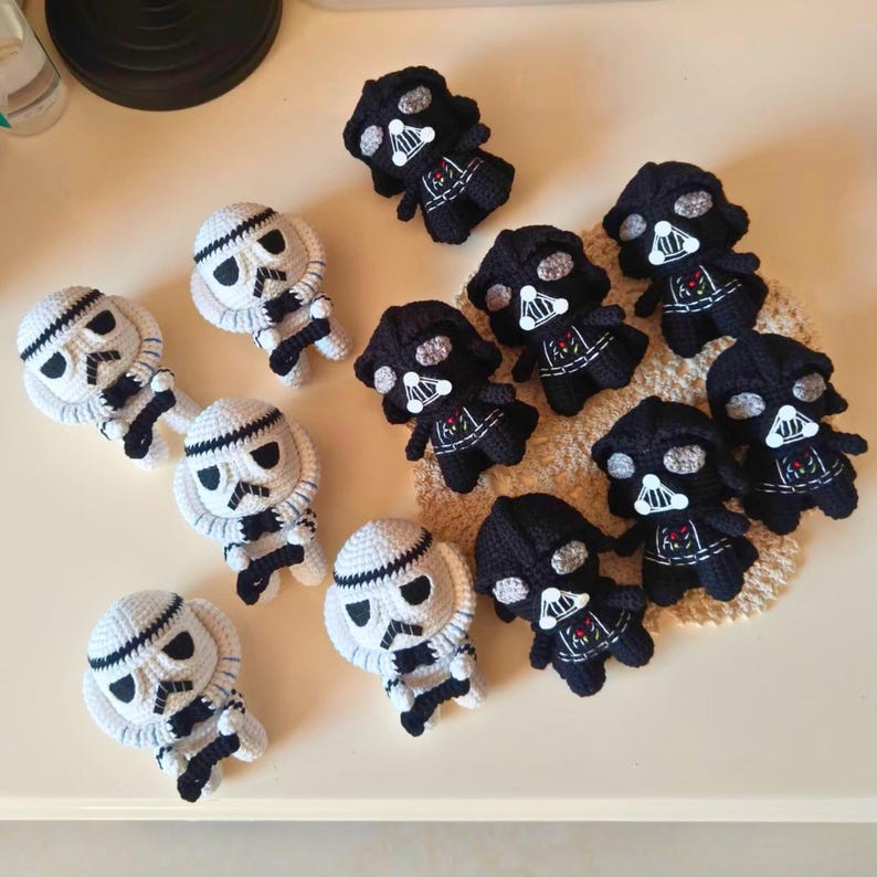 May include: A collection of small, handmade crochet dolls featuring black Darth Vader figures and white Stormtrooper figures. Each character has distinct stitched facial details, including large eyes and mask patterns, crafted with yarn.
