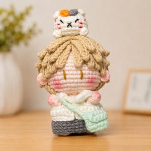 May include: A handmade crochet doll featuring a character with light tan hair, pink cheeks, and a white shirt with a light green crossbody bag over dark gray pants. A small white, orange, and gray cat figure sits atop the character's head.