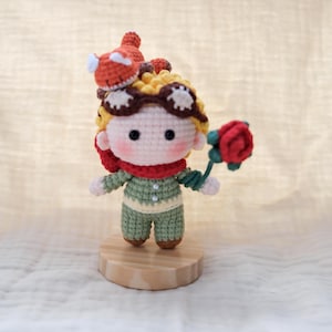 May include: A crocheted doll with yellow hair, wearing brown goggles and a red scarf, dressed in a light green shirt with two white buttons and matching pants. The doll holds a red rose and has a small orange fox figure perched on its head.
