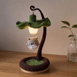Op de afbeelding: Een handgemaakte gehaakte lamp met een groene, bladvormige kap en een bruine steel. Een klein, grijs en wit wezen hangt onder het licht. De basis is bruin met groene accenten.