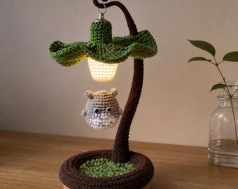 Pode incluir: Uma luminária de crochê artesanal com uma cúpula verde em forma de folha e uma haste marrom. Uma pequena criatura cinza e branca pende sob a luz. A base é marrom com detalhes verdes.
