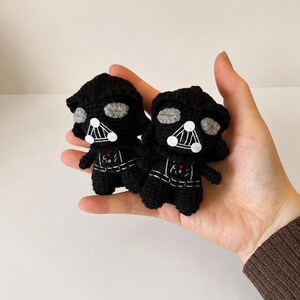 May include: Two small, black crocheted figures featuring a helmeted head with grey eye lenses, a triangular mouth grill with white accents, and a chest panel with small red and green details. These yarn-based dolls are crafted with visible stitches.