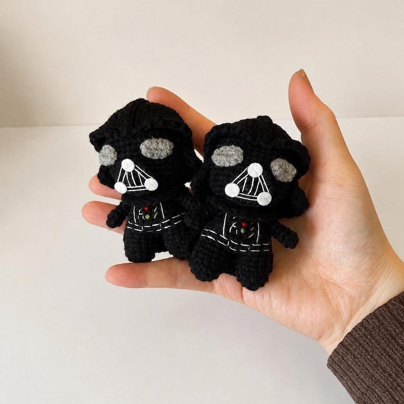 May include: Two small, black crocheted figures featuring a helmeted head with grey eye lenses, a triangular mouth grill with white accents, and a chest panel with small red and green details. These yarn-based dolls are crafted with visible stitches.