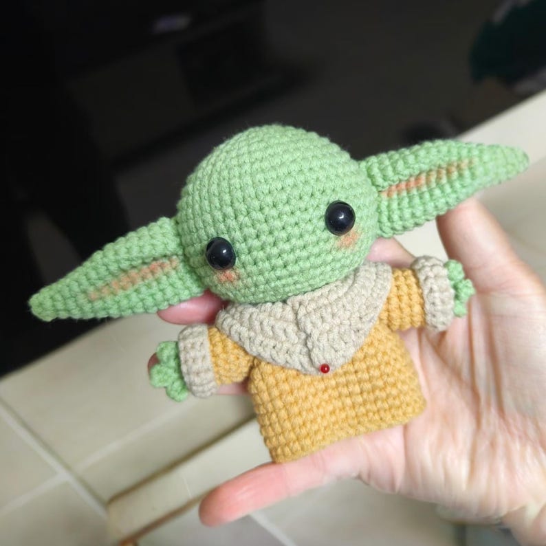 May include: A small, handmade crochet doll featuring a light green head with large black eyes, pointed ears, and pink cheeks. The doll wears a tan-colored robe with a beige collar and has small green hands extending from the sleeves of the garment.