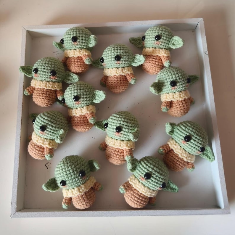 May include: A collection of ten small, handmade crochet figures featuring green heads with large black eyes, pointed ears, and light tan bodies. Each figure is crafted with visible yarn stitches and arranged within a white square tray.