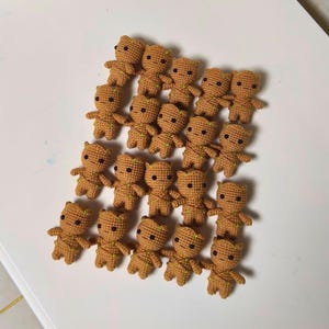 May include: A collection of eighteen small, tan, crocheted figures arranged in a grid. Each figure features a rounded head, a textured body with green yarn accents, and two black bead eyes, resembling a small tree-like character in a uniform style.