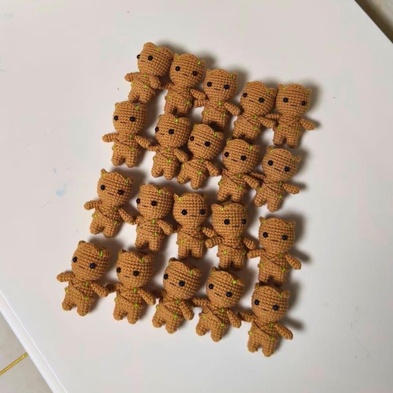 May include: A collection of eighteen small, tan, crocheted figures arranged in a grid. Each figure features a rounded head, a textured body with green yarn accents, and two black bead eyes, resembling a small tree-like character in a uniform style.