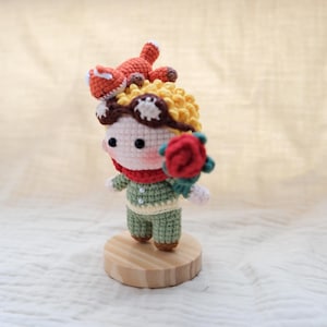 May include: A small crocheted doll with yellow hair, brown goggles, and a red scarf, wearing a green outfit with two white buttons. The doll holds a red rose in one hand and has a small orange fox figure perched on top of its head, all standing on a round wooden base.