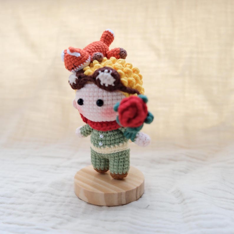May include: A small crocheted doll with yellow hair, brown goggles, and a red scarf, wearing a green outfit with two white buttons. The doll holds a red rose in one hand and has a small orange fox figure perched on top of its head, all standing on a round wooden base.