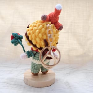 May include: A small crocheted figure with yellow hair, wearing a green outfit, holding a red flower with green leaves. A small orange fox sits on the figure's head. A silver metal keychain ring is attached to the back of the figure's head.