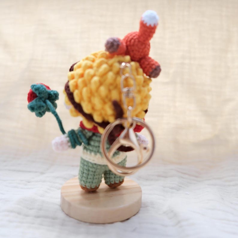May include: A small crocheted figure with yellow hair, wearing a green outfit, holding a red flower with green leaves. A small orange fox sits on the figure's head. A silver metal keychain ring is attached to the back of the figure's head.