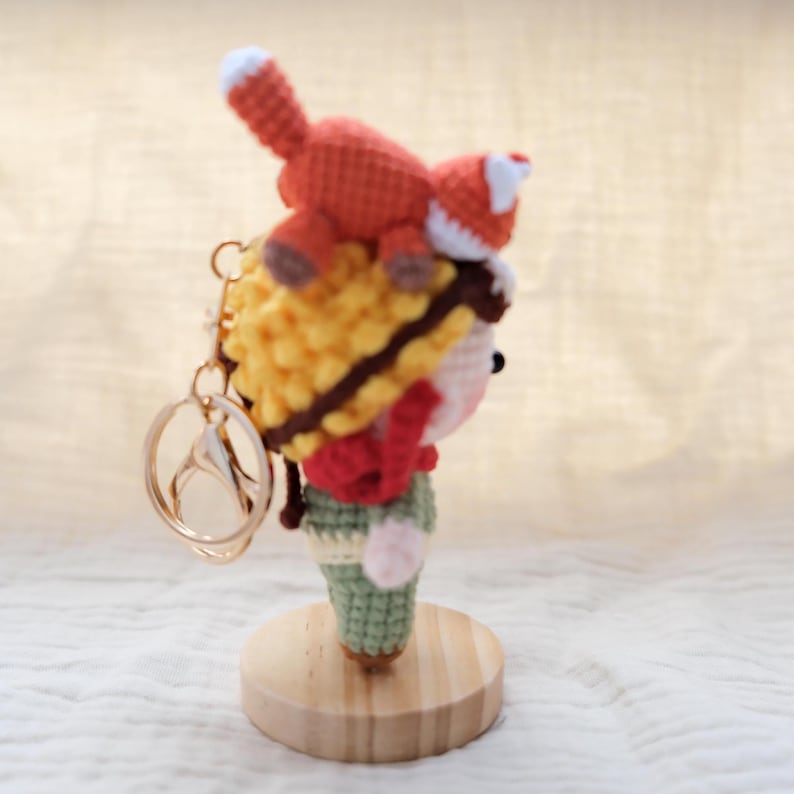 May include: A crochet keychain featuring a small figure with yellow hair and a red scarf, wearing green pants, with a small orange and white fox perched on top of the head. The figure is attached to a gold-toned metal keyring and lobster clasp.