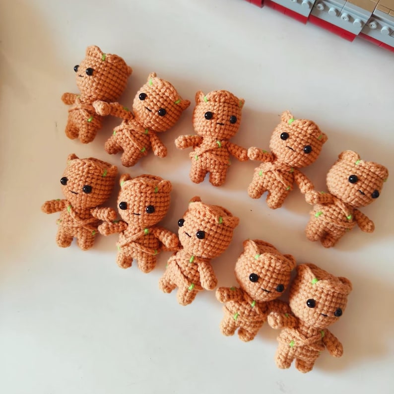 May include: A collection of nine small, light brown crocheted figures, each featuring a textured yarn surface, black bead eyes, and small green yarn accents. The figures are arranged in a loose group, displaying a consistent humanoid shape.