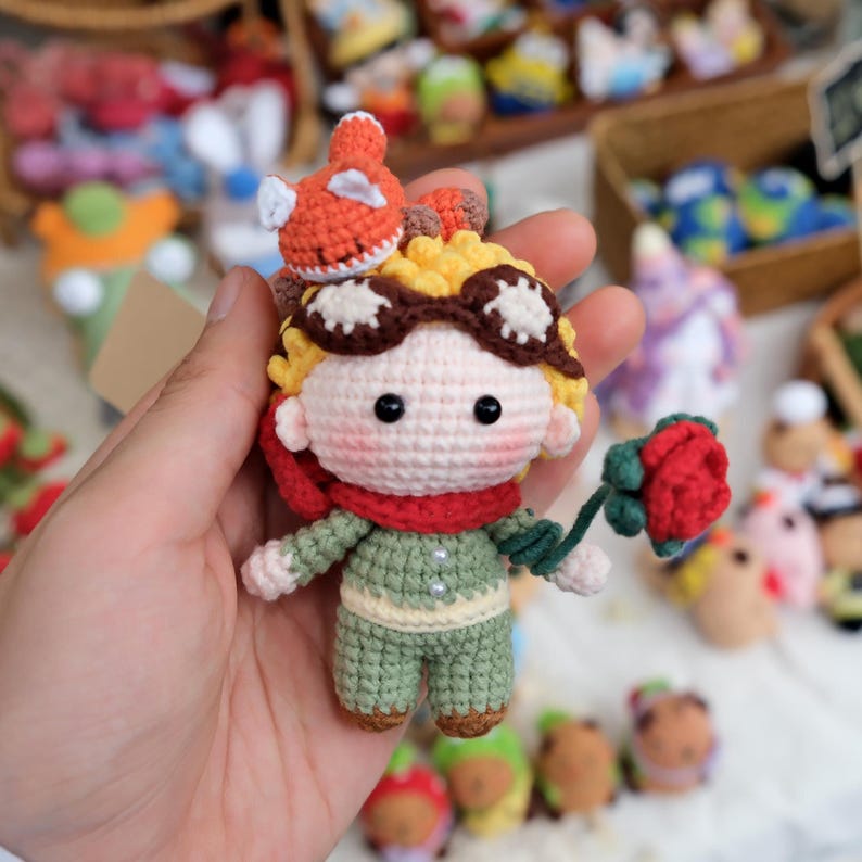 May include: A small crocheted doll with yellow hair, brown goggles, and a red scarf. The doll wears a light green outfit with two white buttons and holds a single red rose with a green stem. An orange fox figure sits atop the doll's head.