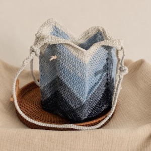 May include: A handmade crochet bucket bag in shades of blue and gray, with a drawstring closure. The bag is placed on a round, woven tray. The bag's design features a chevron pattern, and it has a matching crochet strap.