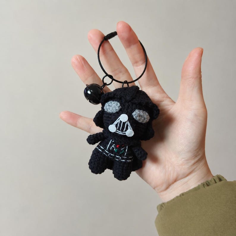 May include: A black crocheted keychain featuring a character with a helmet, gray eye lenses, and a triangular mouth grill. The figure has white and red embroidered details on the chest and is attached to a black circular ring with a small bell.