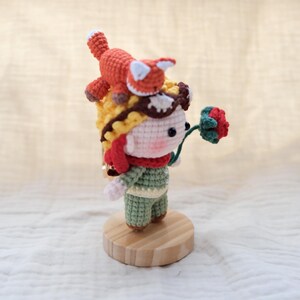 May include: A small crochet doll featuring a pale face with pink cheeks, wearing a yellow hat with brown goggles, a red scarf, and a green outfit. A small orange fox sits on top of the hat, and the doll holds a single red flower with green leaves.