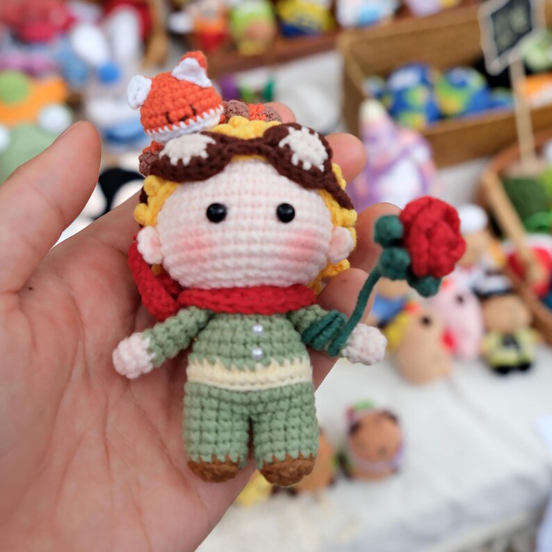 May include: A small crocheted doll featuring a figure with yellow hair, brown goggles, a red scarf, and a green outfit. The doll has a small orange fox perched on its head and is holding a single red rose with a green stem in its hand.