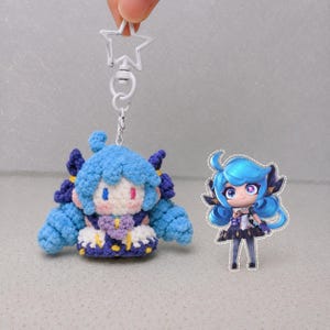 May include: A crochet plush keychain featuring a blue-haired character with a white star-shaped clasp, accompanied by a small acrylic standee of the same character with bright blue hair, large purple eyes, and a dark outfit with gold accents.