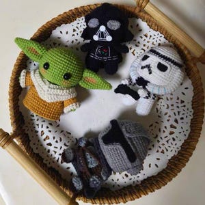 May include: Four crocheted figures including a green creature with large ears and a tan robe, a black helmeted figure, a white armored figure with black accents, and a grey armored figure, all arranged on a white lace doily inside a round wicker tray.