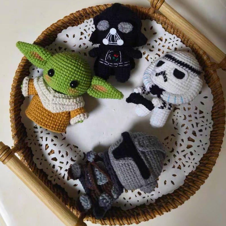 May include: Four crocheted figures including a green creature with large ears and a tan robe, a black helmeted figure, a white armored figure with black accents, and a grey armored figure, all arranged on a white lace doily inside a round wicker tray.