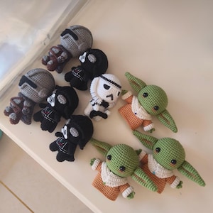 May include: A collection of eight small crocheted figures featuring characters from Star Wars. The set includes two grey armored figures, three black helmeted figures, one white helmeted figure, and two green creatures with large ears and tan robes.
