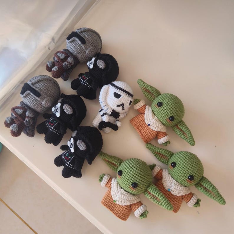 May include: A collection of eight small crocheted figures featuring characters from Star Wars. The set includes two grey armored figures, three black helmeted figures, one white helmeted figure, and two green creatures with large ears and tan robes.