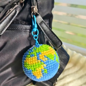 Puede incluir: Un llavero de globo de ganchillo con detalles azules, verdes y amarillos. El dije en forma de Tierra está sujeto a un cierre azul y cuelga de una bolsa negra. Los continentes están representados en verde y amarillo.