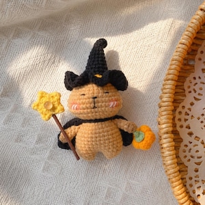 May include: A tan crocheted animal figure wearing a black pointed witch hat and a black cape. The figure has pink cheeks, holds a yellow star-shaped wand with a clear bead in the center, and carries a small orange pumpkin in its other hand.