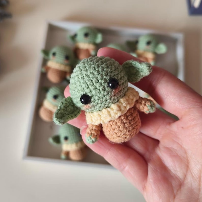 May include: A small, handmade crochet figure featuring a light green head with large black eyes, pointed ears, and a tan-colored body with a cream-colored collar. The figure is crafted with visible yarn stitches and has a soft, rounded appearance.