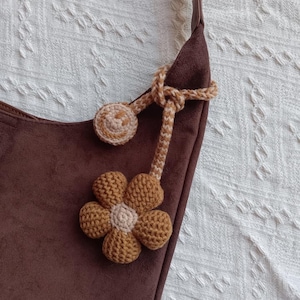 Op de afbeelding: Een donkerbruine suède schoudertas met een decoratief gehaakt accessoire aan de riem. Het accessoire bestaat uit een lichtbruine en beige gehaakte bloem en een kleine ronde gehaakte smiley, verbonden door een gevlochten koord.