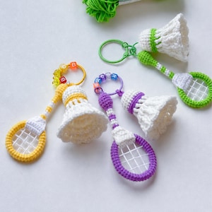 Può includere: Tre portachiavi all'uncinetto fatti a mano che raffigurano racchette da badminton e volani in miniatura. Ogni set include una racchetta con rete bianca, un volano bianco e un anello colorato decorato con piccole perline a lettera gialle, viola e verdi.
