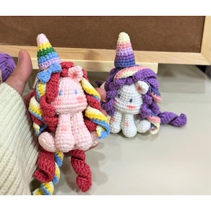 May include: Two handmade crochet unicorn dolls with spiral manes and horns. One unicorn is light pink with a rainbow horn and mane, while the other is white with a purple and yellow horn and mane. Both feature small embroidered eyes and blush.