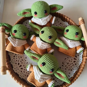 May include: Five small, green, crocheted figures with large black eyes, pink cheeks, and pointed ears. Each figure wears a tan-colored tunic with a cream-colored collar. The figures are arranged together in a circular, woven, brown wicker tray.