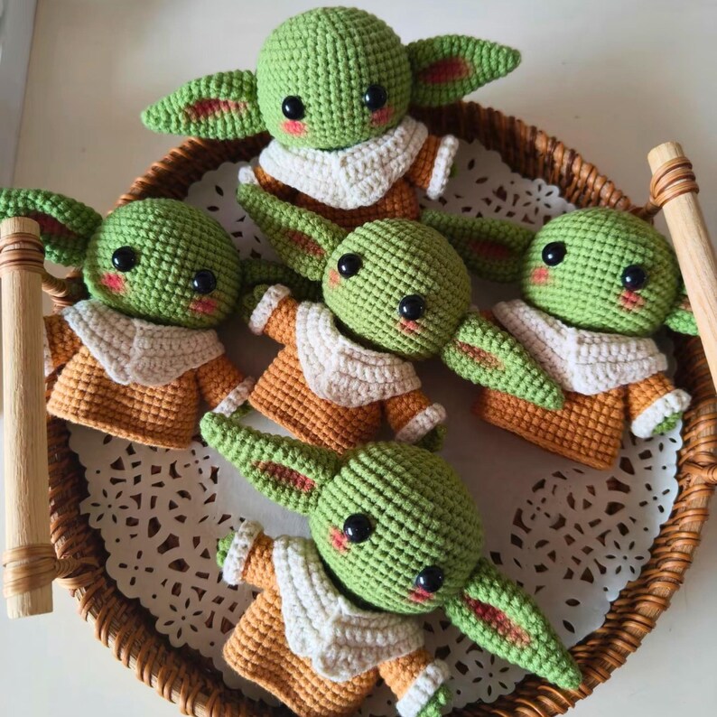 May include: Five small, green, crocheted figures with large black eyes, pink cheeks, and pointed ears. Each figure wears a tan-colored tunic with a cream-colored collar. The figures are arranged together in a circular, woven, brown wicker tray.