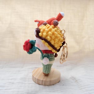 May include: A crochet keychain featuring a small figure with yellow curly hair, a red scarf, and green pants, holding a red flower. An orange fox sits atop the figure's head. The keychain is attached to a gold-toned metal ring and stands on a round wooden base.